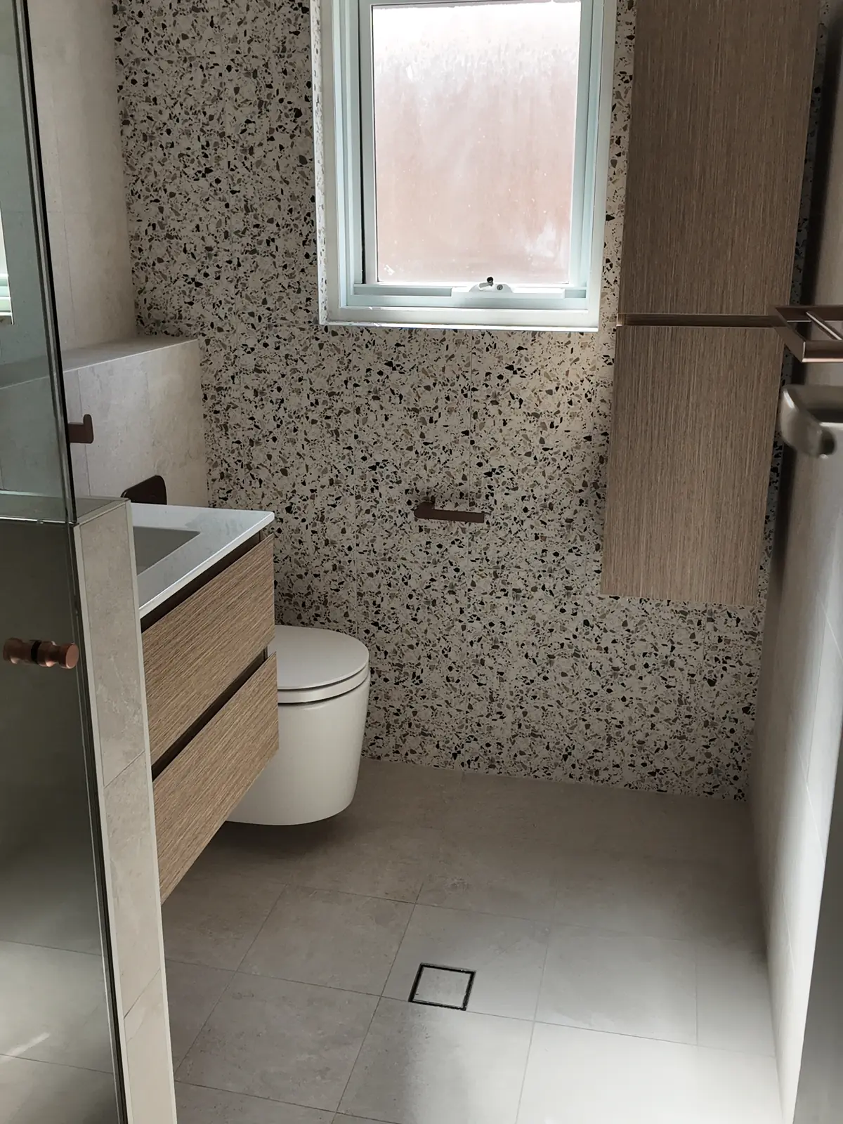 The renovated bathroom from the shower, with a terrazzo-look feature wall, wall-hung vanity and large-format floor tiles