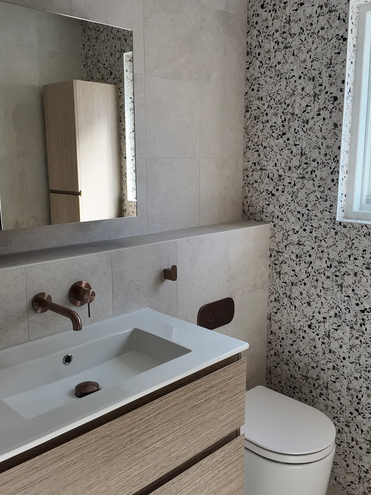 The renovated bathroom looking towards the vanity, with a large mirror, wall-hung storage and the terrazzo-look feature wall