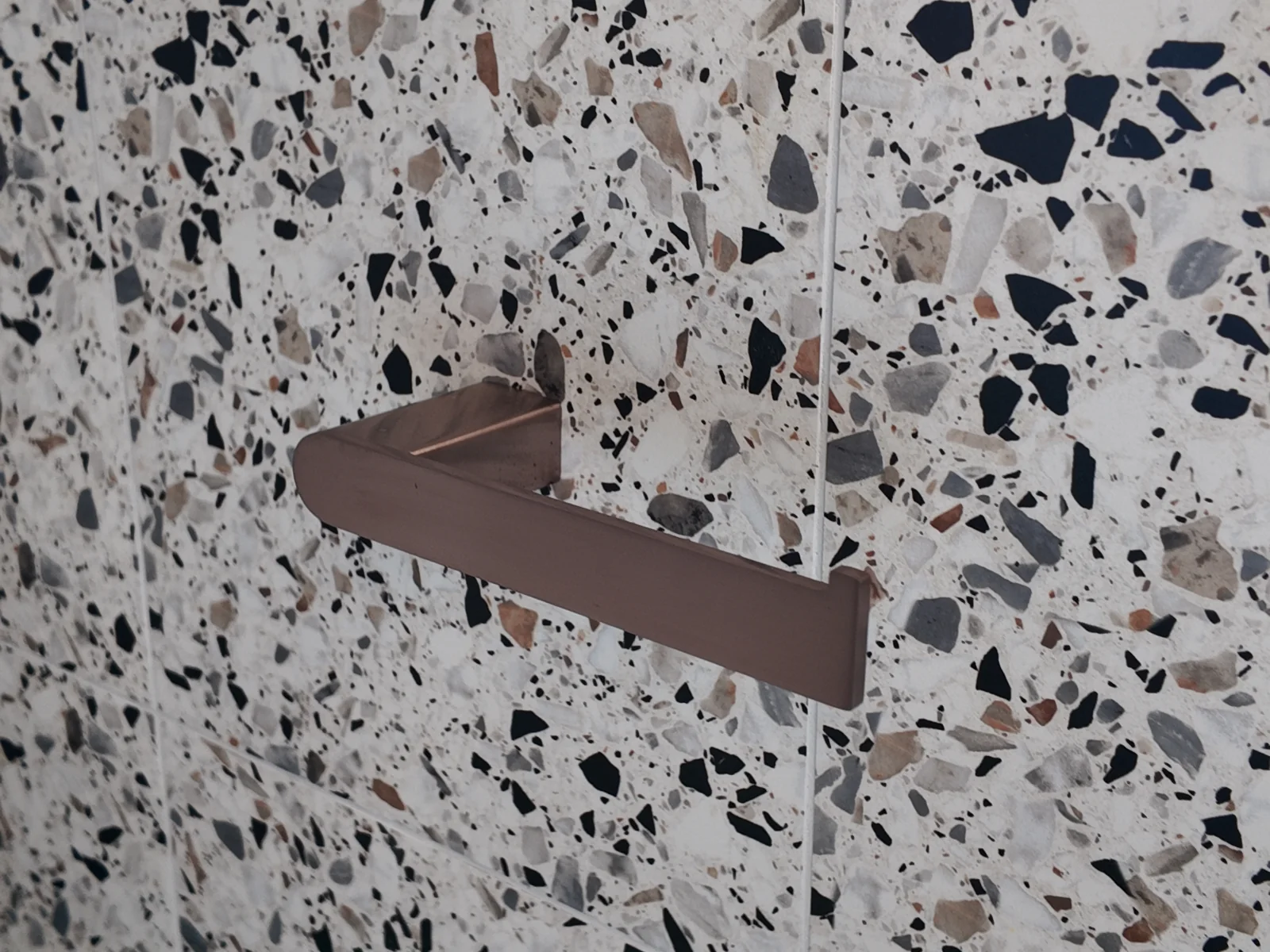 Close-up of the Piura Road terrazzo-look feature tile in gravel matt, with a brushed bronze toilet roll holder