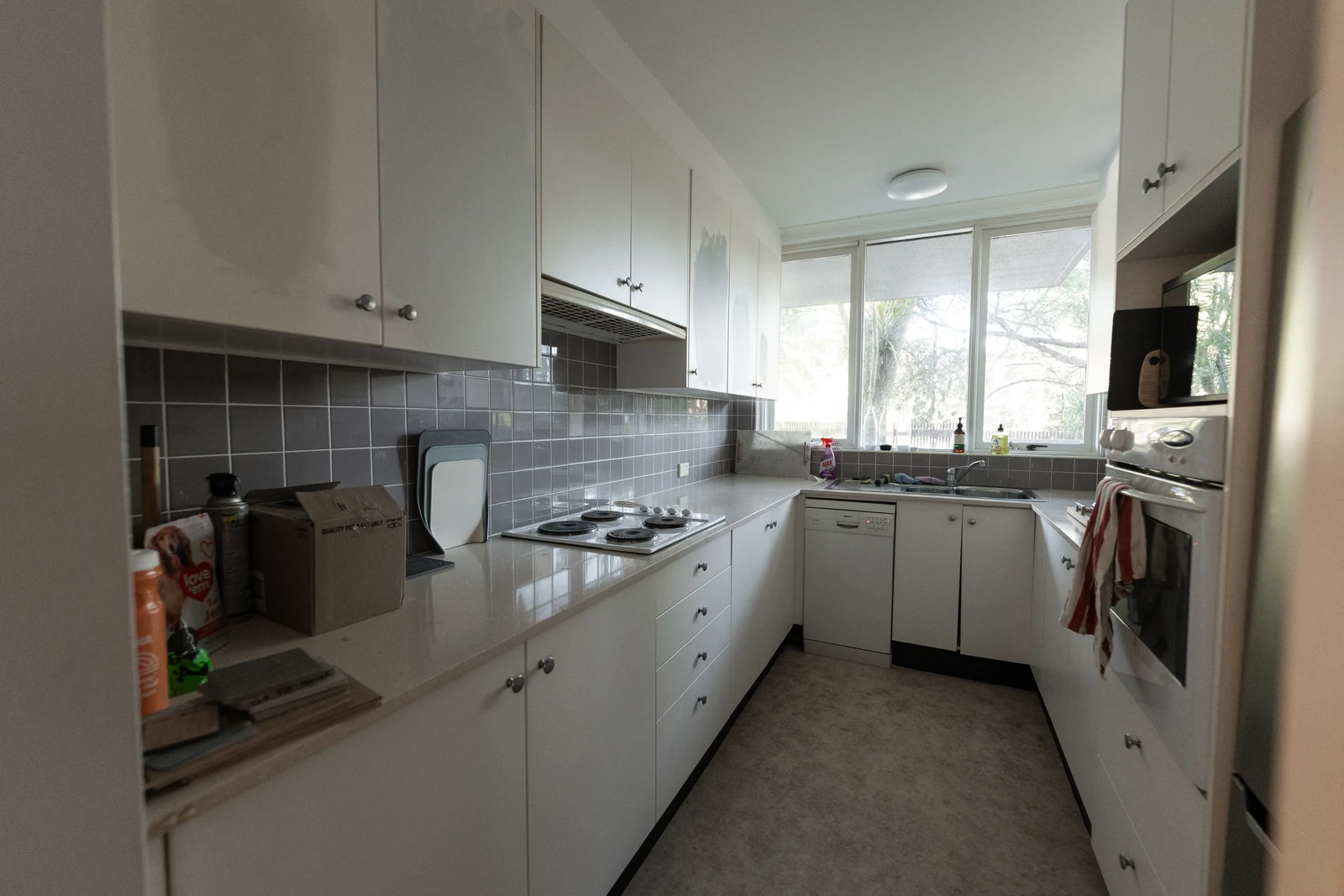 The original galley kitchen before renovation, with white laminate cabinetry, grey tiled splashback and vinyl flooring
